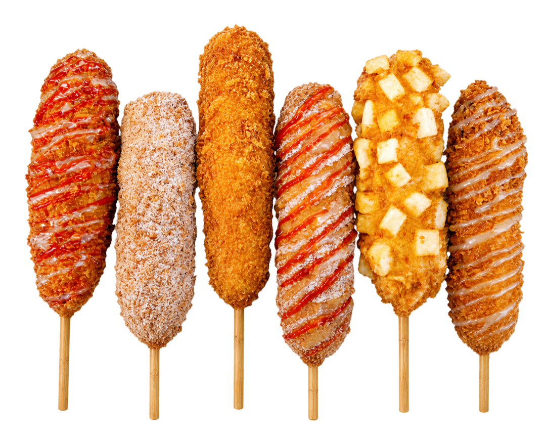 Six Korean corn dogs on sticks with sugar and breadcrumb coatings, ketchup and sauce drizzles, and one covered in potato cubes